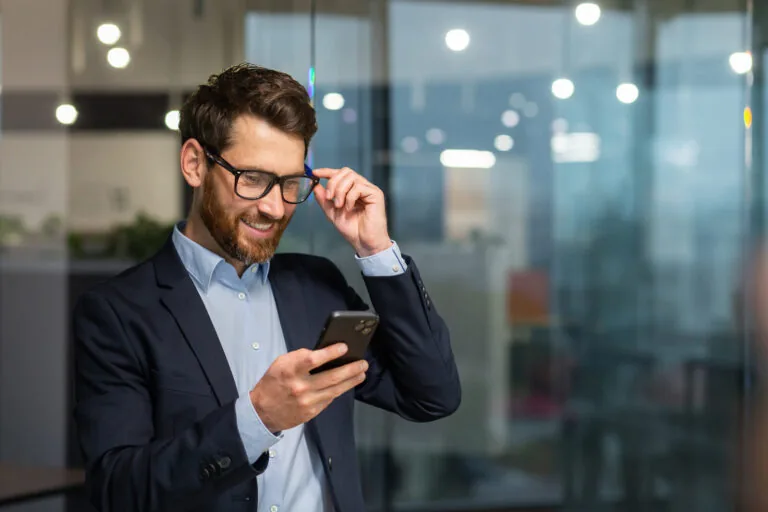 Commercial en costume consultant son smartphone dans un bureau vitré pour structurer son plan de prospection commerciale